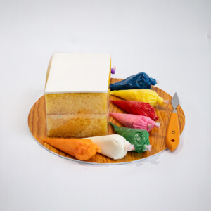 Square cake dessert with white frosting on a wooden tray, surrounded by assorted colourful frostings in a plastic bags and a small knife.