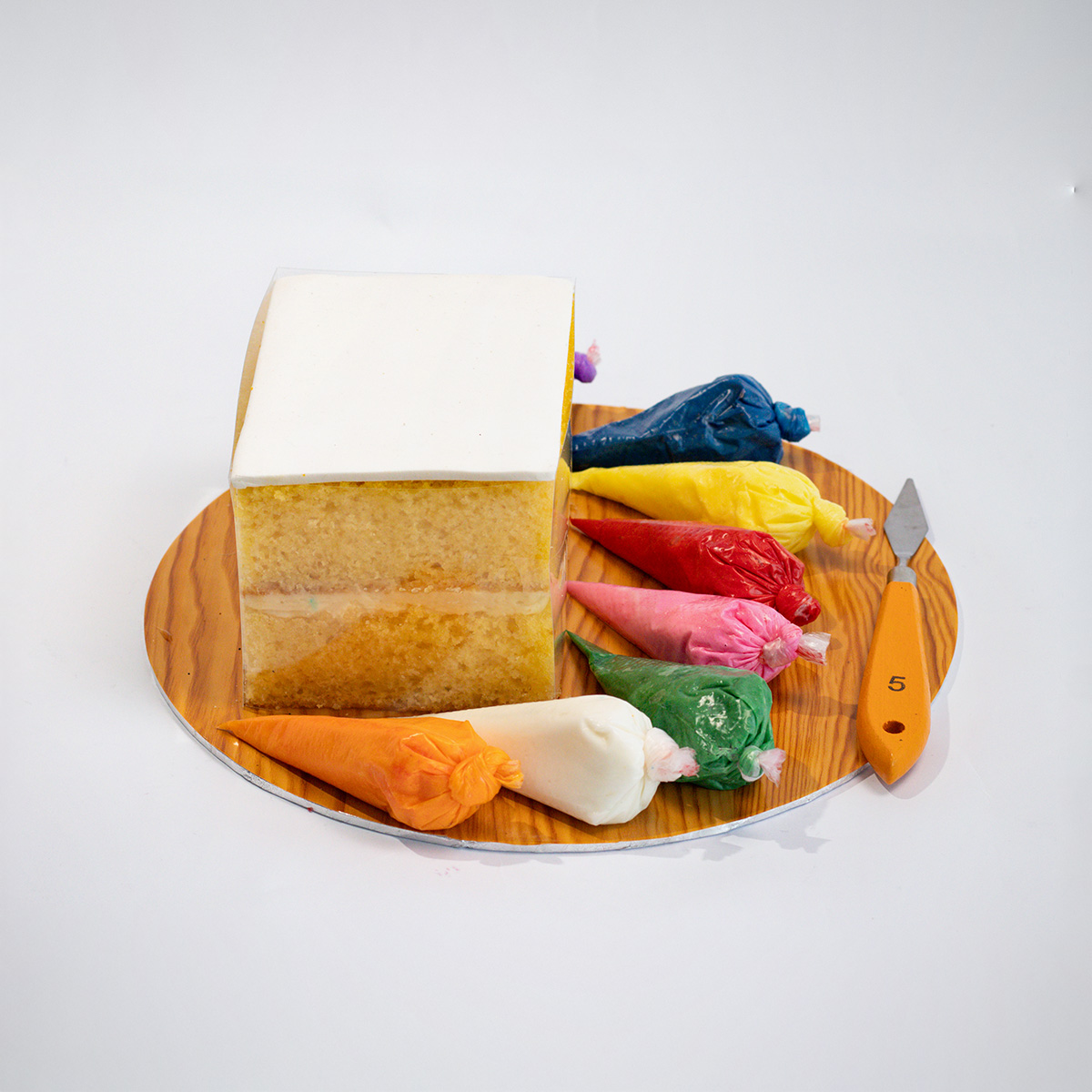 Square cake dessert with white frosting on a wooden tray, surrounded by assorted colourful frostings in a plastic bags and a small knife.