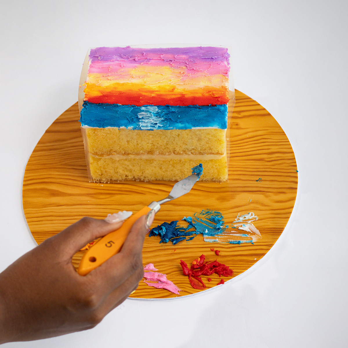 Colourful layered cake with purple, orange, yellow, and blue frosting sections, with a hand actively layering the frosting on a wooden surface.