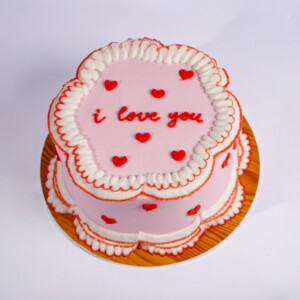 Kiss and Tell lunchbox cake with pink frosting, white piped edges, and “i love you” text in red icing surrounded by small red hearts.
