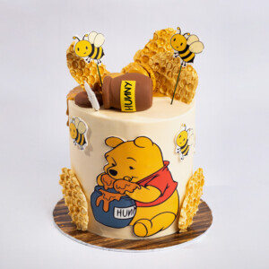 Winnie Inspired Cake featuring Winnie the Pooh holding a pot of honey, surrounded by bees and honeycomb decorations with “HUNNY” text on the pot.