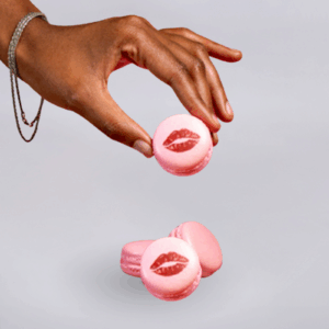One Kiss macaron with pink shell and red lip print design, held in a hand adorned with bracelets on a plain light-coloured background.