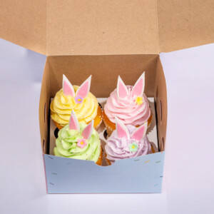 Easter bunny cupcakes in a cardboard box with window, featuring cupcakes topped with yellow, pink, and green frosting and small pink bunny ear decorations.