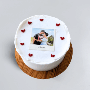 15 cm Picture Perfect cake with white frosting and red heart decorations around the edge, featuring a polaroid-style photo of a couple embracing in the centre and the text “I love you” below the image.
