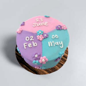 A pastel three-tone buttercream celebration cake divided into pink, lavender, and teal sections, decorated with small floral accents and personalised with the dates “23 June,” “02 Feb,” and “06 May” written on top.